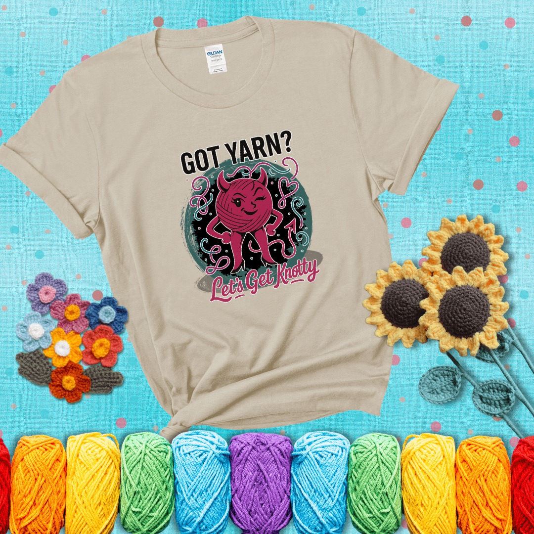 A cheeky pink yarn ball with devil horns, arms, and legs winks under the text “Got Yarn? Let’s Get Knotty,” showing a beige tee with a teal sunflower background
