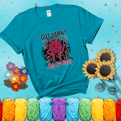 A cheeky pink yarn ball with devil horns, arms, and legs winks under the text “Got Yarn? Let’s Get Knotty,” showing a blue-green tee with a teal sunflower background
