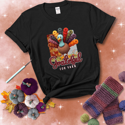 A crocheted turkey with colorful yarn skeins for tail feathers, standing on fall leaves with the text “Grateful for Yarn” showing a black tee with a festive Fall background