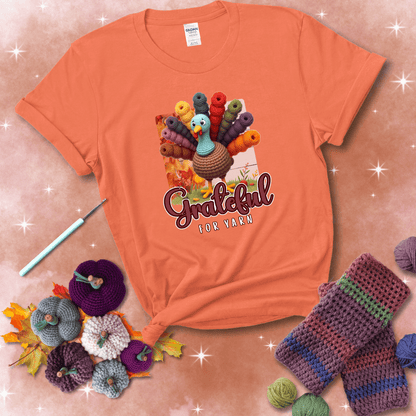 A crocheted turkey with colorful yarn skeins for tail feathers, standing on fall leaves with the text “Grateful for Yarn” showing a light orange tee with a festive Fall background