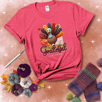 A crocheted turkey with colorful yarn skeins for tail feathers, standing on fall leaves with the text “Grateful for Yarn” showing a light red tee with a festive Fall background