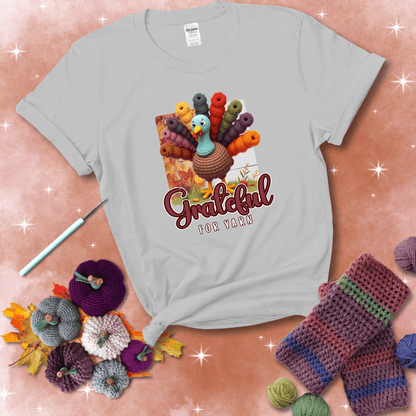 A crocheted turkey with colorful yarn skeins for tail feathers, standing on fall leaves with the text “Grateful for Yarn” showing a light grey tee with a festive Fall background