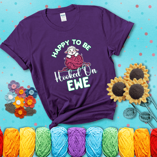 Smiling cartoon sheep peeks from a pink yarn ball, surrounded by green text “Happy to Be Hooked on Ewe” — playful and affectionate sheep pun for crocheters showing a purple tee with a teal sunflower background