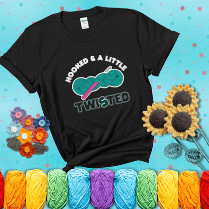 A teal skein of yarn with a hot pink crochet hook through it, captioned “Hooked & a Little Twisted” showing a black tee with a teal sunflower background