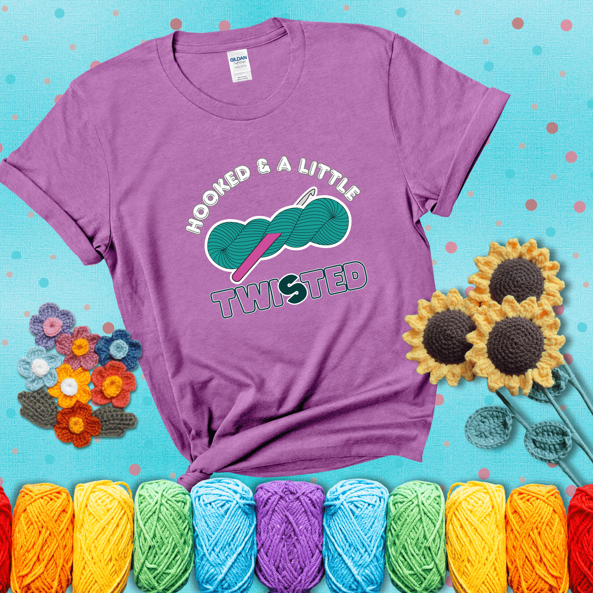 A teal skein of yarn with a hot pink crochet hook through it, captioned “Hooked & a Little Twisted” showing a light purple tee with a teal sunflower background