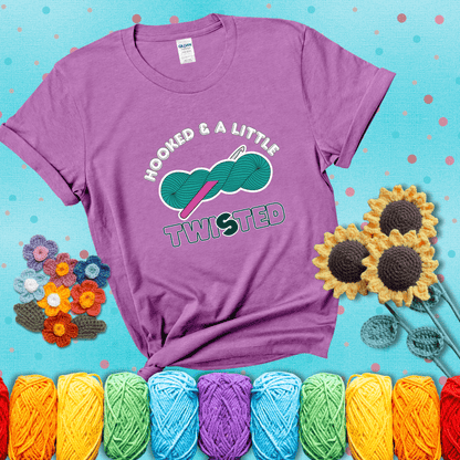 A teal skein of yarn with a hot pink crochet hook through it, captioned “Hooked & a Little Twisted” showing a light purple tee with a teal sunflower background
