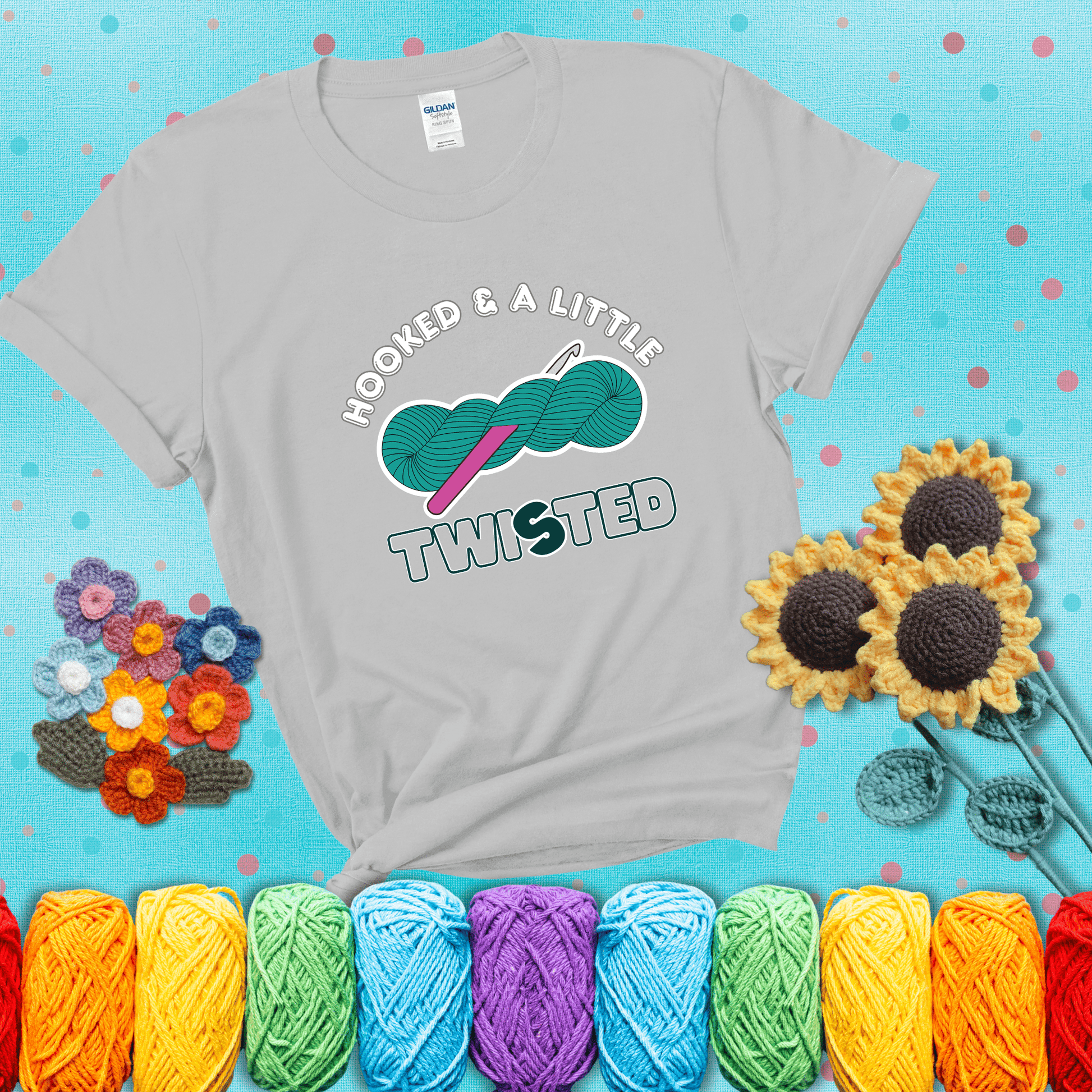 A teal skein of yarn with a hot pink crochet hook through it, captioned “Hooked & a Little Twisted” showing a light grey tee with a teal sunflower background