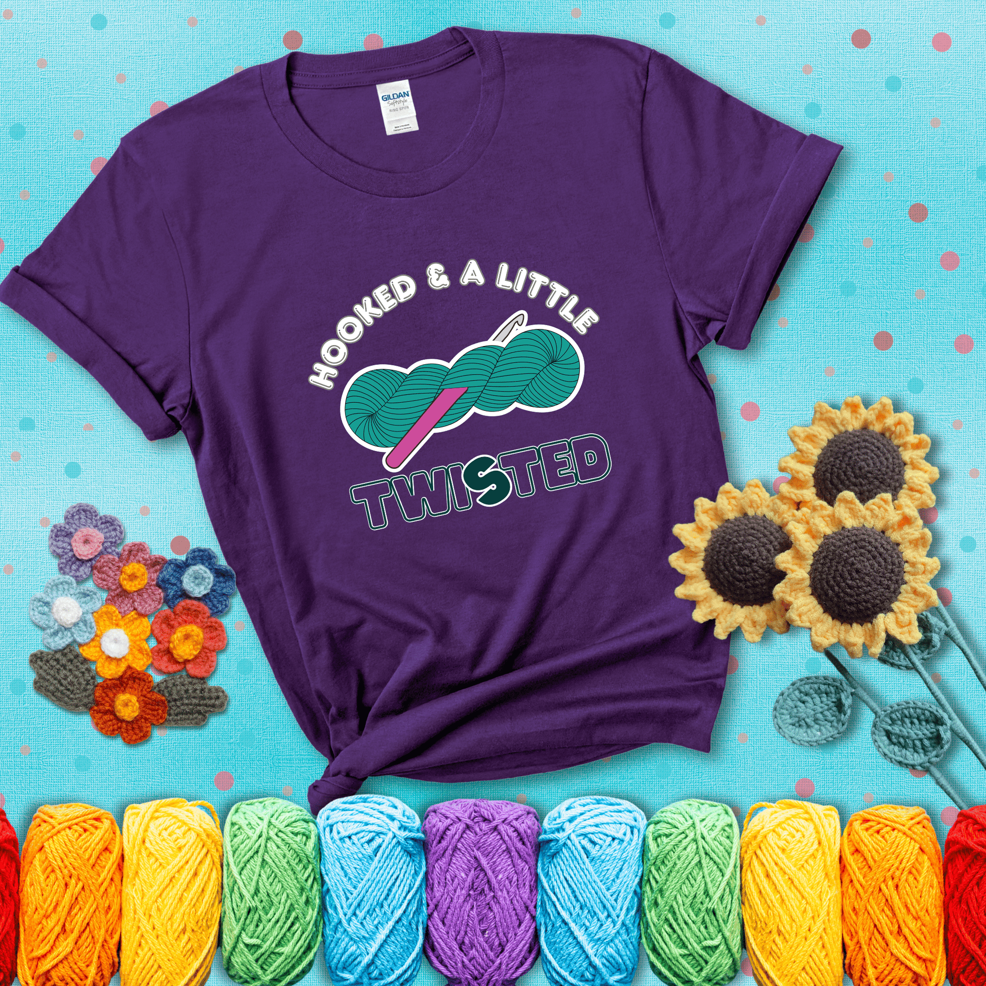 A teal skein of yarn with a hot pink crochet hook through it, captioned “Hooked & a Little Twisted” showing a purple tee with a teal sunflower background