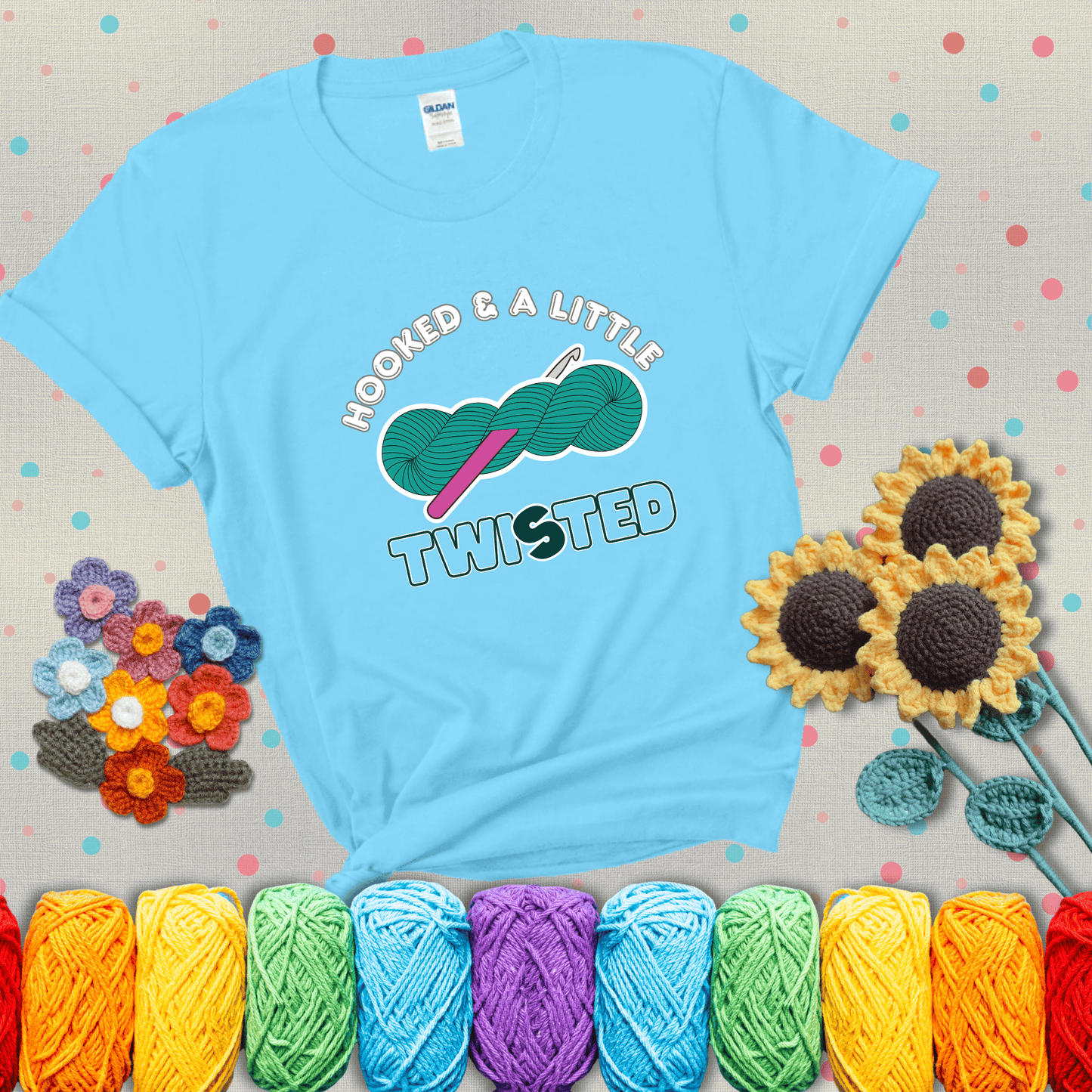 A teal skein of yarn with a hot pink crochet hook through it, captioned “Hooked & a Little Twisted” showing a light blue tee with grey sunflower background