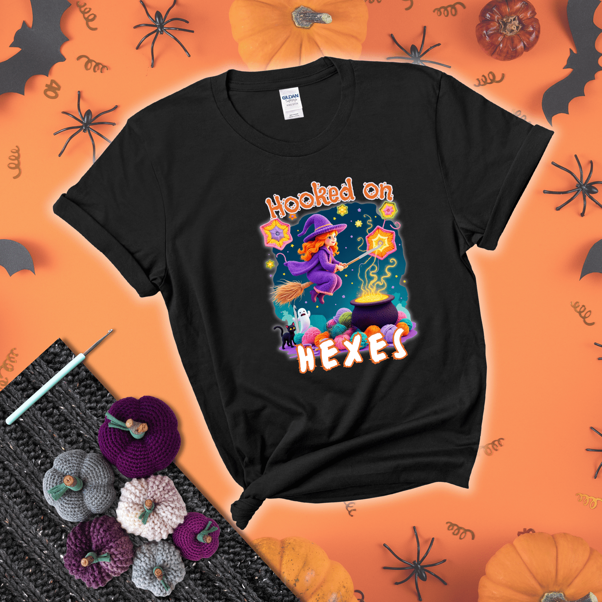 Witchy vibe with stars, hexagons, and a crochet hook. Text reads: “Hooked on Hexes.” A mystical and moody tee for fans of spooky stitches showing a black tee with a festive Halloween background