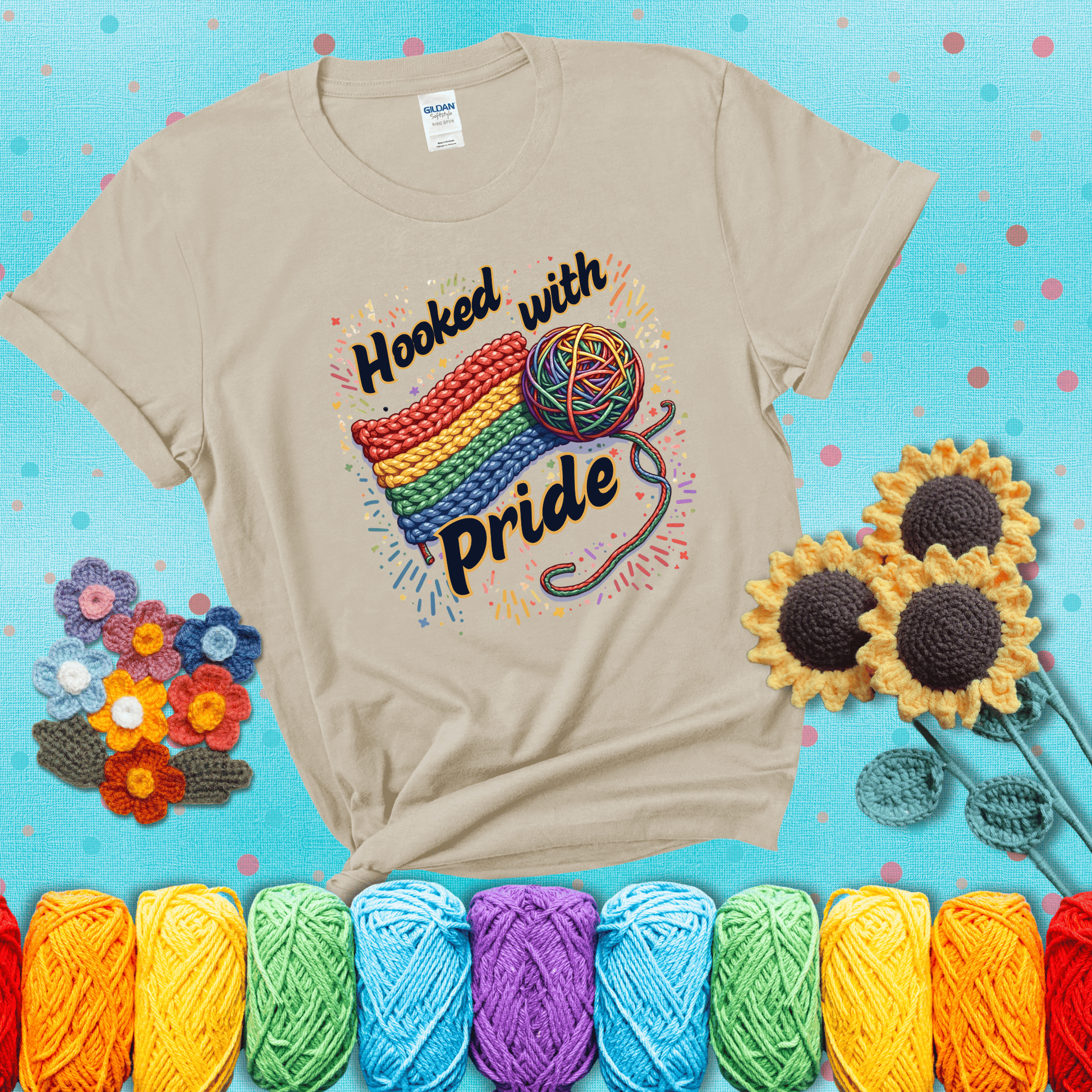 A rainbow-striped crochet swatch paired with a multicolor yarn ball and bold “Hooked With Pride” lettering. Confetti surrounds the vibrant scene showing a beige tee with a teal sunflower background