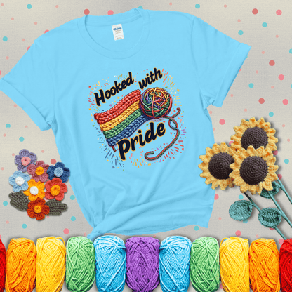 A rainbow-striped crochet swatch paired with a multicolor yarn ball and bold “Hooked With Pride” lettering. Confetti surrounds the vibrant scene showing a light blue tee with grey sunflower background