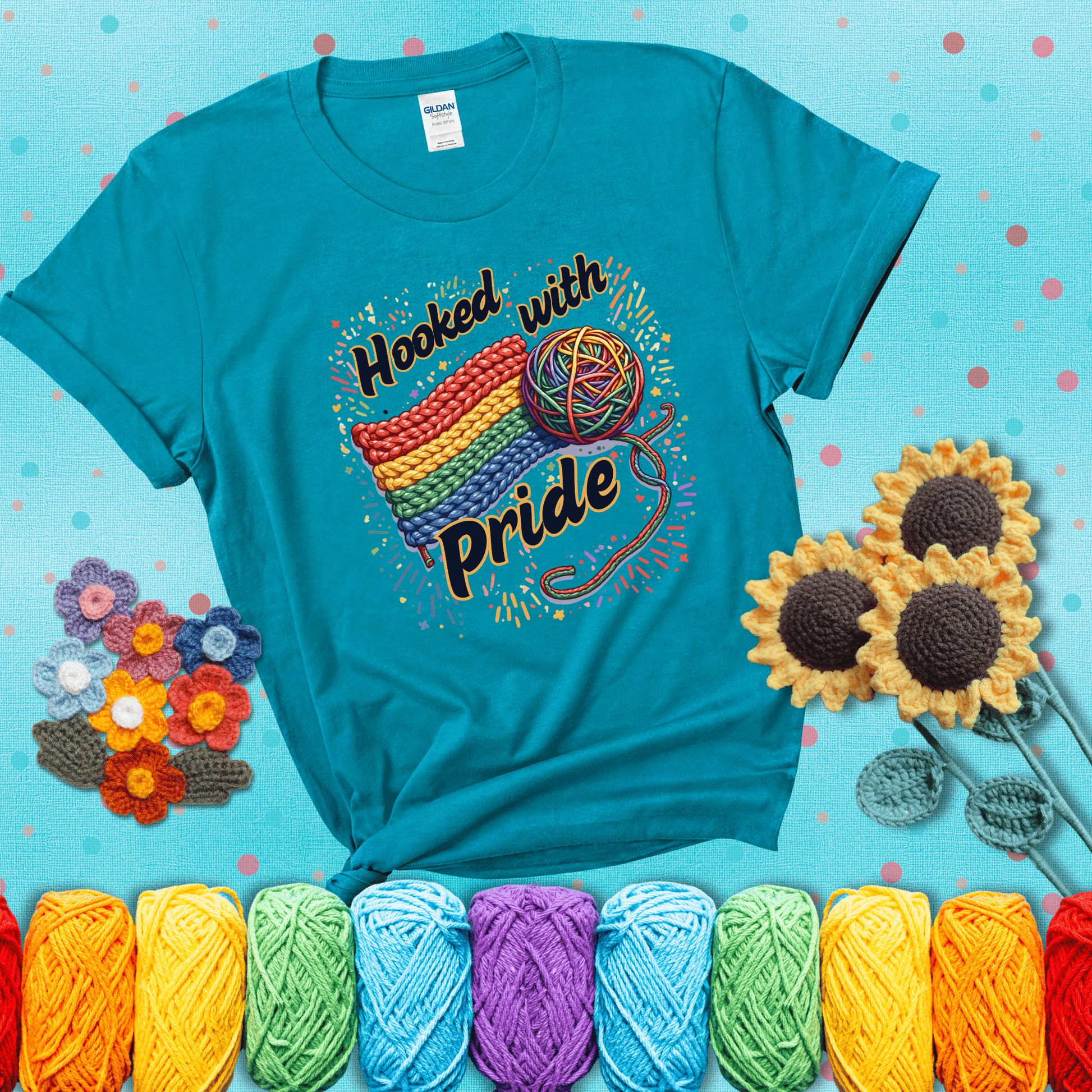 A rainbow-striped crochet swatch paired with a multicolor yarn ball and bold “Hooked With Pride” lettering. Confetti surrounds the vibrant scene showing a blue-green tee with a teal sunflower background