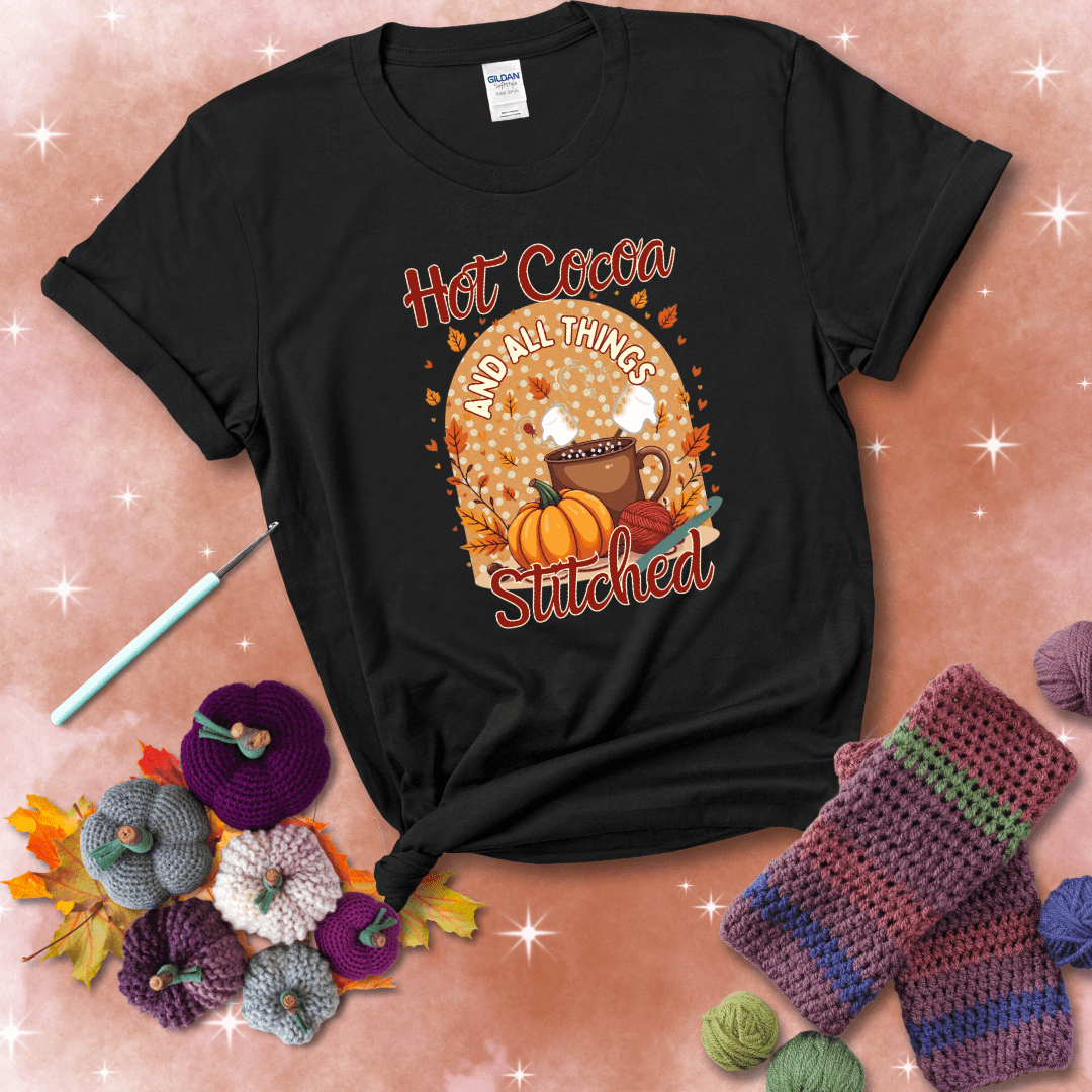 A cozy fall scene with a cup of hot cocoa topped with marshmallows, a pumpkin, yarn balls, and autumn leaves. The phrase reads “Hot Cocoa and All Things Stitched.” showing a black tee with a festive Fall background
