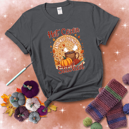 A cozy fall scene with a cup of hot cocoa topped with marshmallows, a pumpkin, yarn balls, and autumn leaves. The phrase reads “Hot Cocoa and All Things Stitched.” showing a dark grey tee with a festive Fall background