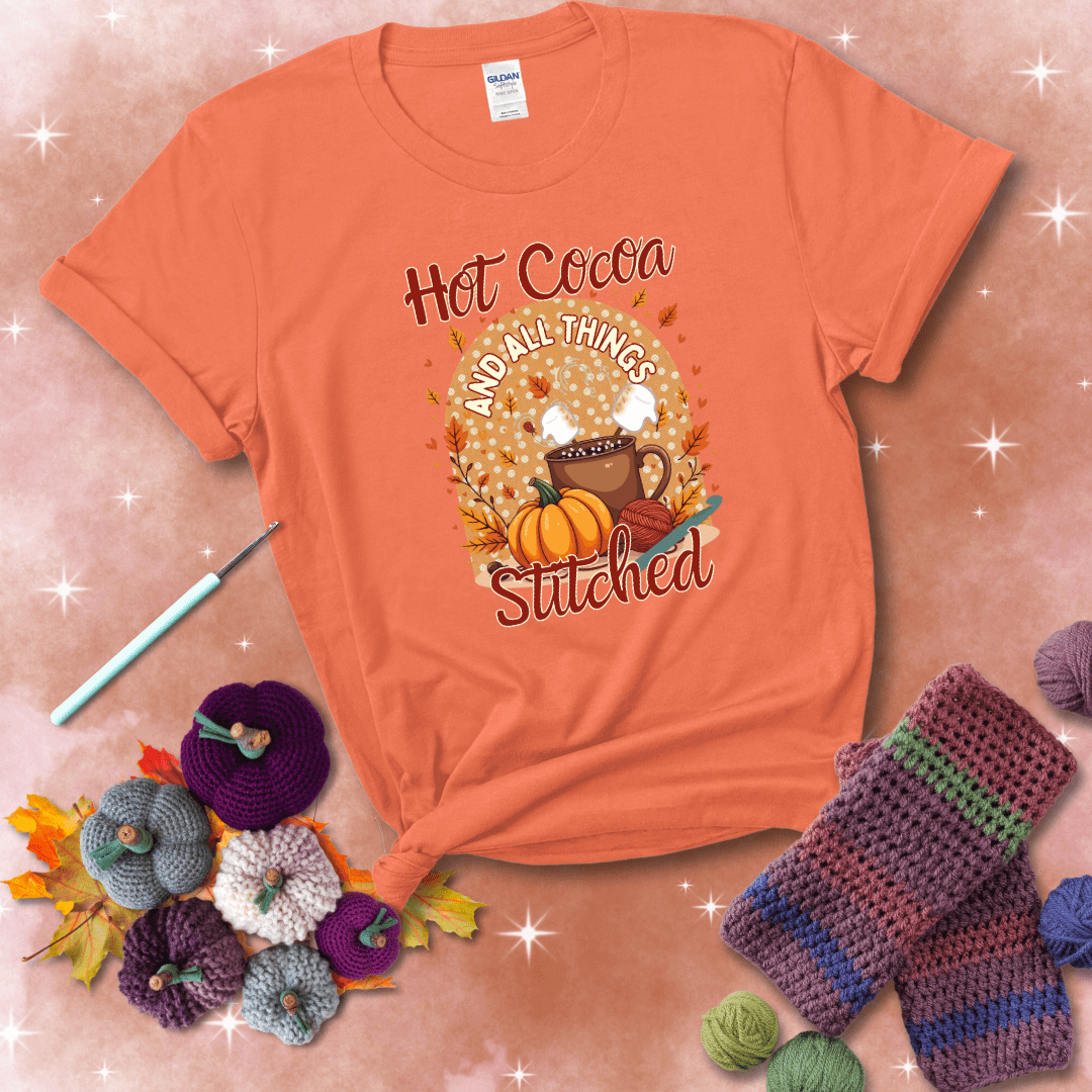 A cozy fall scene with a cup of hot cocoa topped with marshmallows, a pumpkin, yarn balls, and autumn leaves. The phrase reads “Hot Cocoa and All Things Stitched.” showing a light orange tee with a festive Fall background