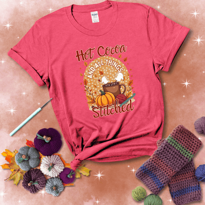 A cozy fall scene with a cup of hot cocoa topped with marshmallows, a pumpkin, yarn balls, and autumn leaves. The phrase reads “Hot Cocoa and All Things Stitched.” showing a light red tee with a festive Fall background