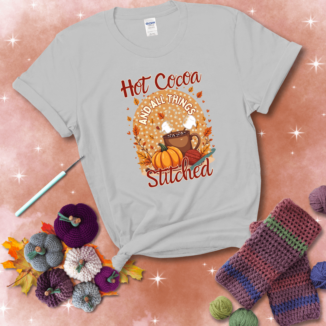 A cozy fall scene with a cup of hot cocoa topped with marshmallows, a pumpkin, yarn balls, and autumn leaves. The phrase reads “Hot Cocoa and All Things Stitched.” showing a light grey tee with a festive Fall background
