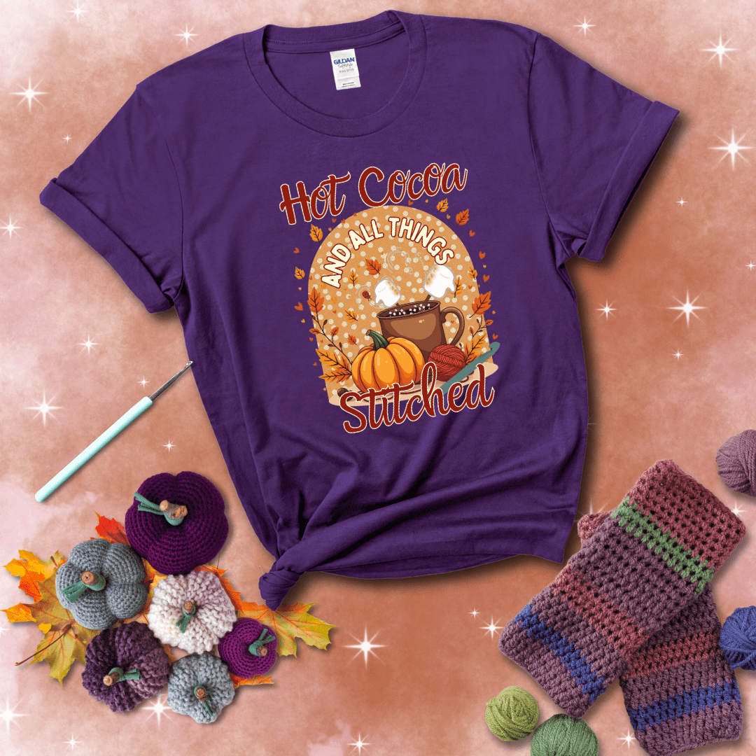 A cozy fall scene with a cup of hot cocoa topped with marshmallows, a pumpkin, yarn balls, and autumn leaves. The phrase reads “Hot Cocoa and All Things Stitched.” showing a purple tee with a festive Fall background