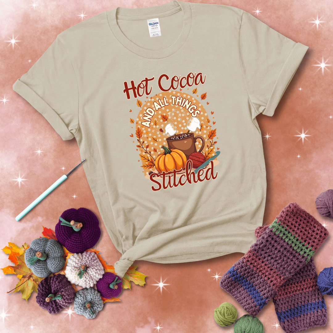 A cozy fall scene with a cup of hot cocoa topped with marshmallows, a pumpkin, yarn balls, and autumn leaves. The phrase reads “Hot Cocoa and All Things Stitched.” showing a beige tee with a festive Fall background