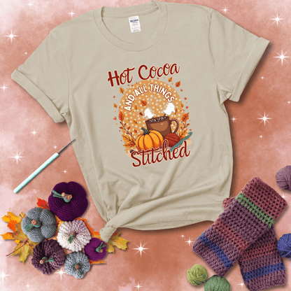 A cozy fall scene with a cup of hot cocoa topped with marshmallows, a pumpkin, yarn balls, and autumn leaves. The phrase reads “Hot Cocoa and All Things Stitched.” showing a beige tee with a festive Fall background