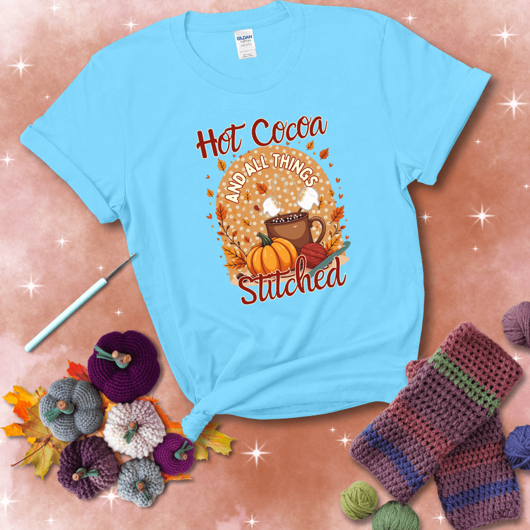 A cozy fall scene with a cup of hot cocoa topped with marshmallows, a pumpkin, yarn balls, and autumn leaves. The phrase reads “Hot Cocoa and All Things Stitched.” showing a light blue tee with a festive Fall background