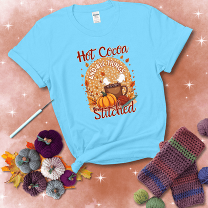 A cozy fall scene with a cup of hot cocoa topped with marshmallows, a pumpkin, yarn balls, and autumn leaves. The phrase reads “Hot Cocoa and All Things Stitched.” showing a light blue tee with a festive Fall background