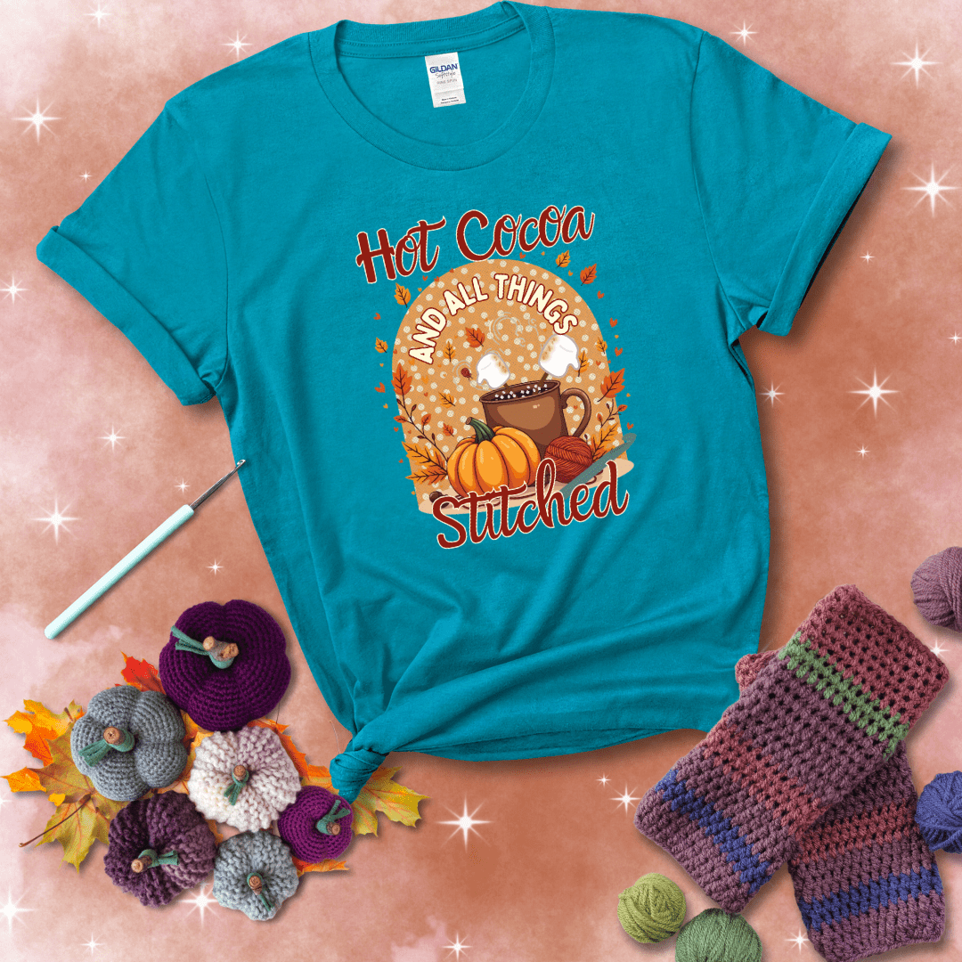 A cozy fall scene with a cup of hot cocoa topped with marshmallows, a pumpkin, yarn balls, and autumn leaves. The phrase reads “Hot Cocoa and All Things Stitched.” showing a blue-green tee with a festive Fall background