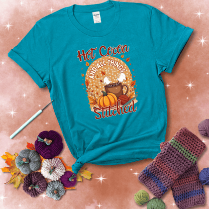 A cozy fall scene with a cup of hot cocoa topped with marshmallows, a pumpkin, yarn balls, and autumn leaves. The phrase reads “Hot Cocoa and All Things Stitched.” showing a blue-green tee with a festive Fall background