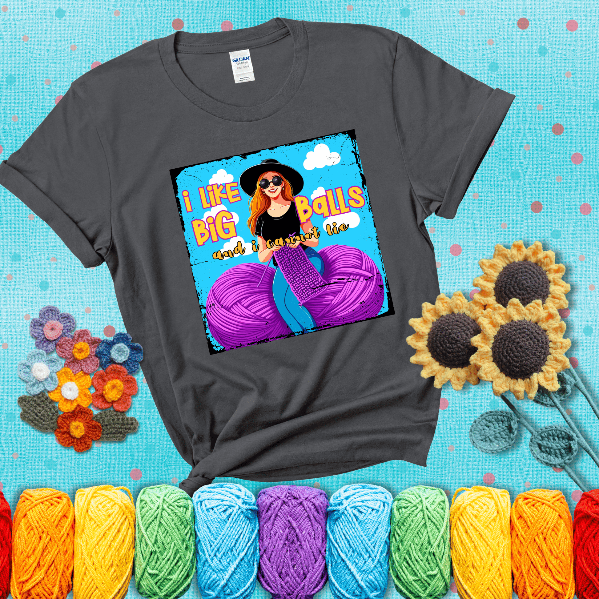 Bold cartoon image of a woman crocheting between two giant purple yarn balls with the phrase “I like big balls and I cannot lie” in retro fonts showing a dark grey tee with a teal sunflower background