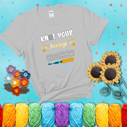 Bold lettering with a yarn hook graphic. Text reads: “I'm Not Your Average Hooker.” A cheeky crochet tee for confident crafters who own their skills, showing a light grey tee with a teal sunflower background