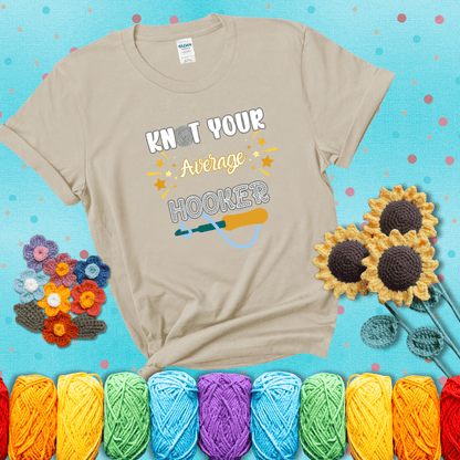 Bold lettering with a yarn hook graphic. Text reads: “I'm Not Your Average Hooker.” A cheeky crochet tee for confident crafters who own their skills, showing a beige tee with a teal sunflower background