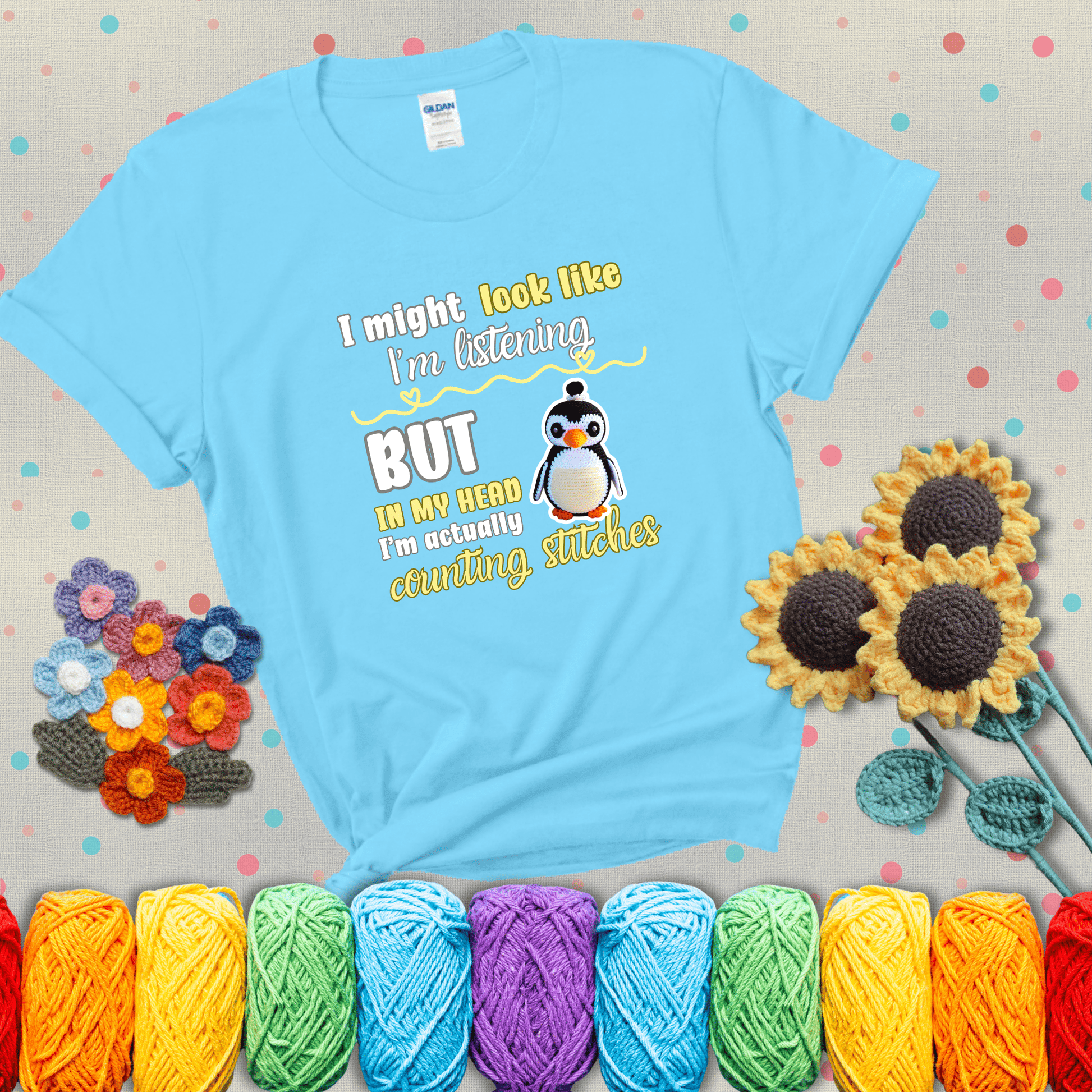 Crochet penguin next to the text “I might look like I’m listening but in my head I’m actually counting stitches” showing a light blue tee with grey sunflower background