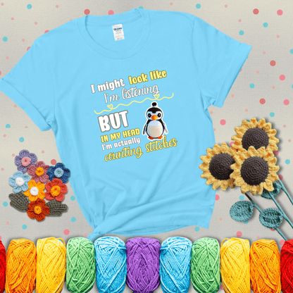 Crochet penguin next to the text “I might look like I’m listening but in my head I’m actually counting stitches” showing a light blue tee with grey sunflower background