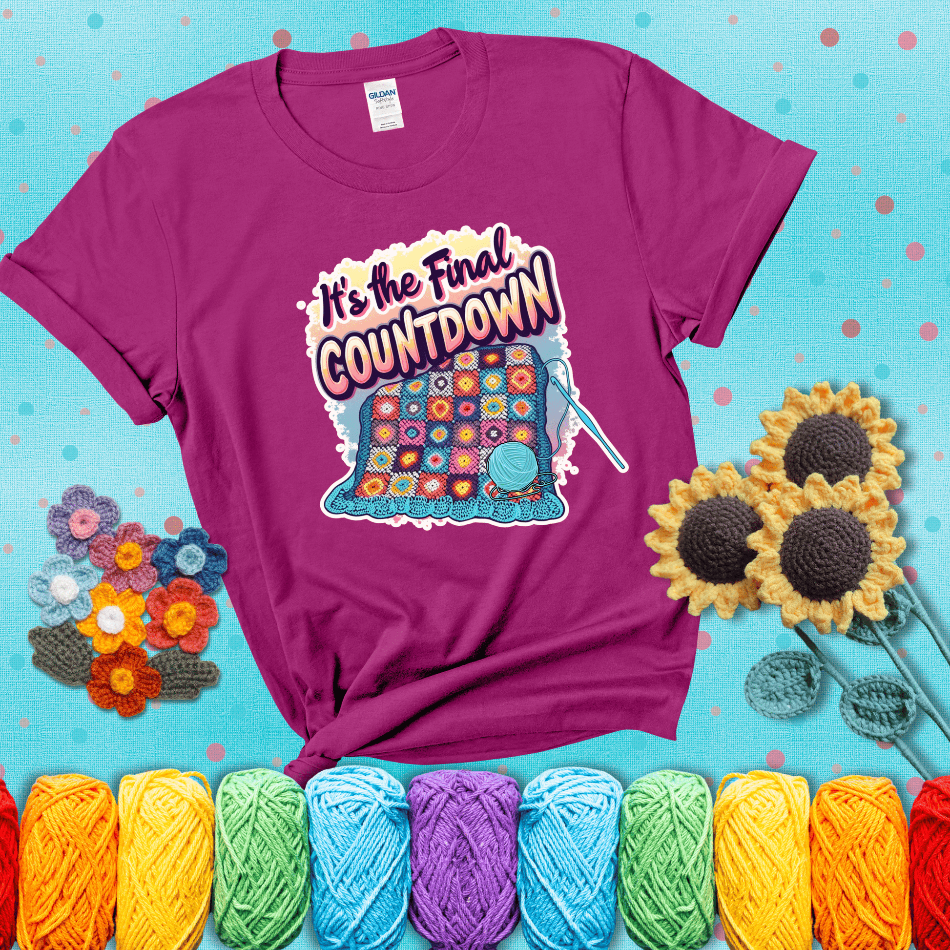 Colorful crochet blanket in progress with text "It’s the Final Countdown" above it in bold, multicolor lettering. A blue ball of yarn and hook are placed at the bottom, showing a magenta tee with a teal sunflower background