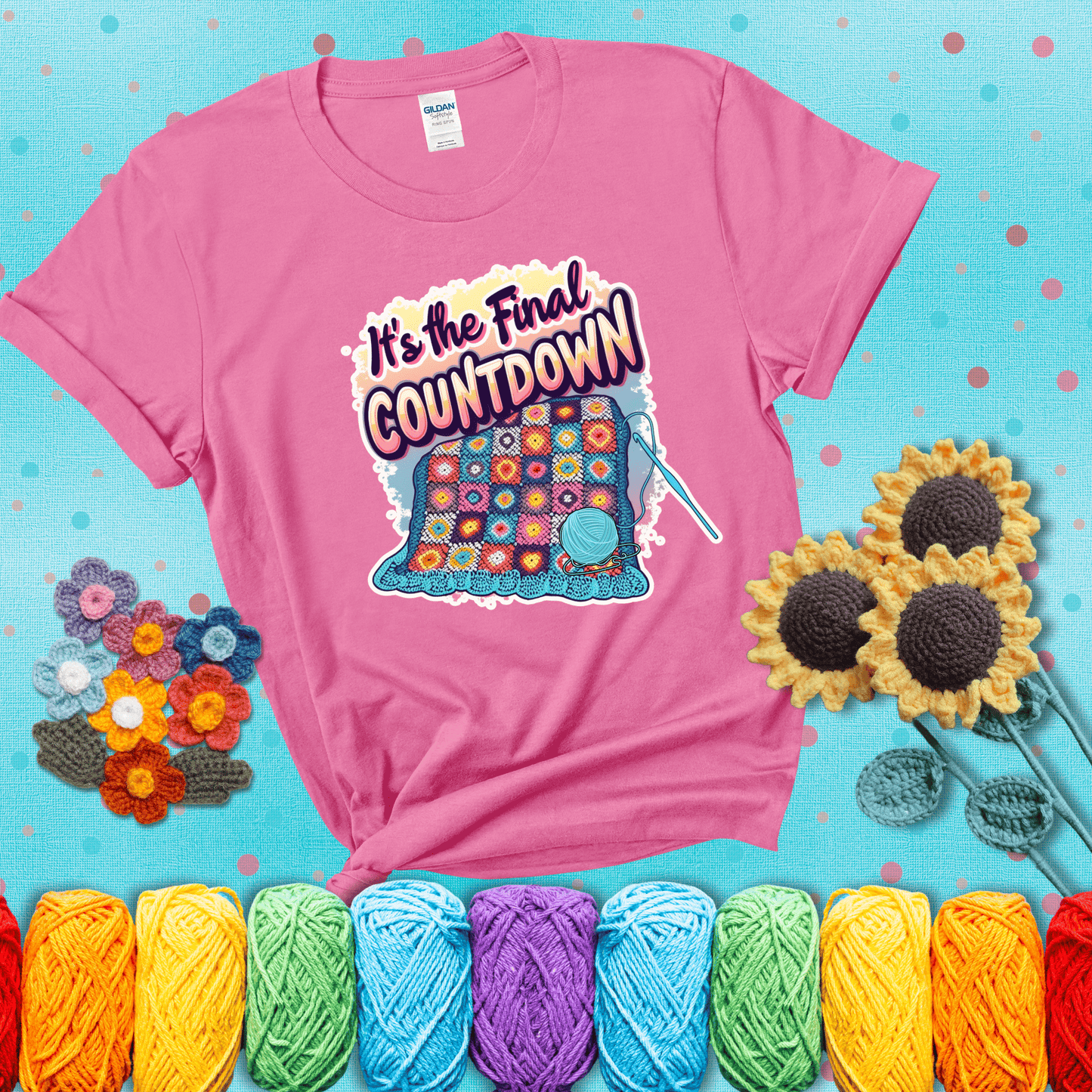 Colorful crochet blanket in progress with text "It’s the Final Countdown" above it in bold, multicolor lettering. A blue ball of yarn and hook are placed at the bottom, showing a pink tee with a teal sunflower background