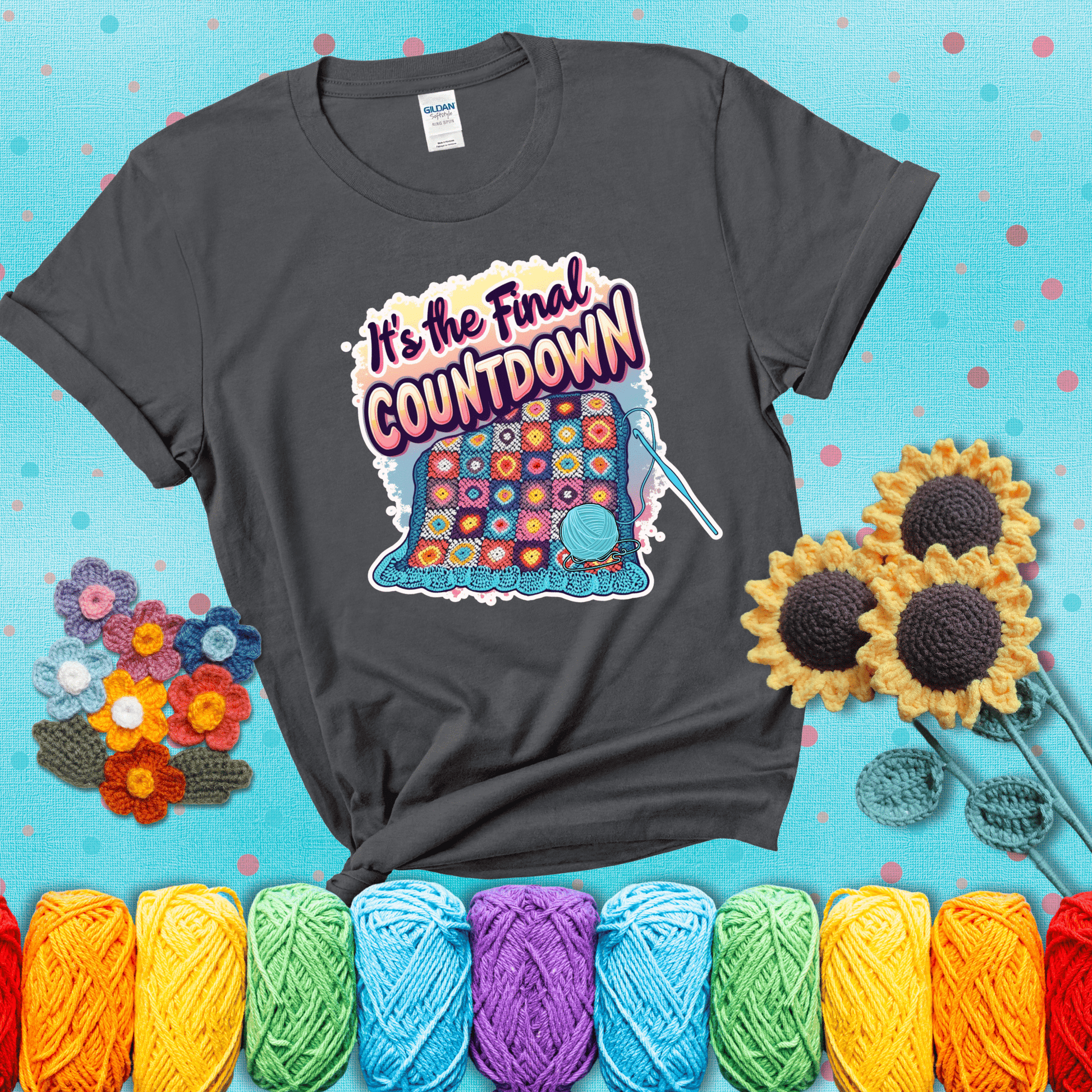 Colorful crochet blanket in progress with text "It’s the Final Countdown" above it in bold, multicolor lettering. A blue ball of yarn and hook are placed at the bottom, showing a dark grey tee with a teal sunflower background