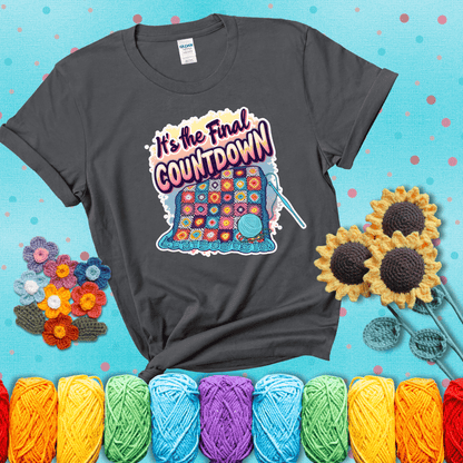 Colorful crochet blanket in progress with text "It’s the Final Countdown" above it in bold, multicolor lettering. A blue ball of yarn and hook are placed at the bottom, showing a dark grey tee with a teal sunflower background