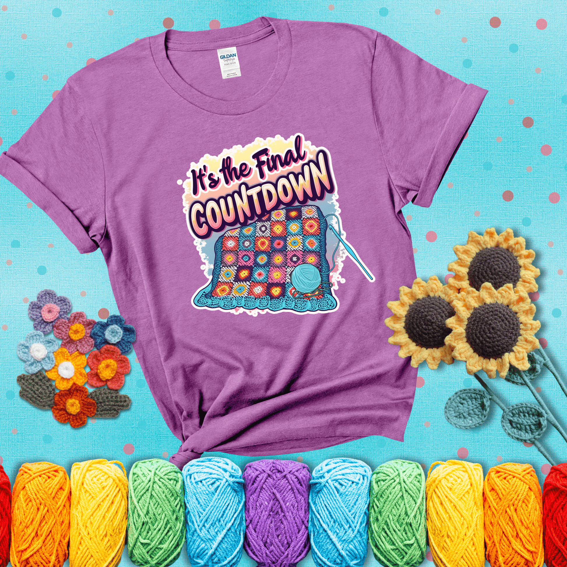 Colorful crochet blanket in progress with text "It’s the Final Countdown" above it in bold, multicolor lettering. A blue ball of yarn and hook are placed at the bottom, showing a light purple tee with a teal sunflower background
