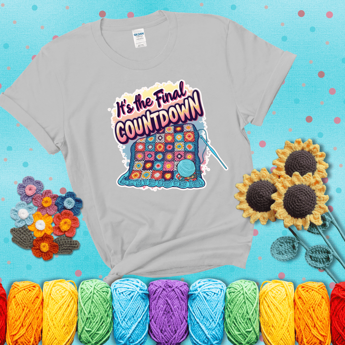 Colorful crochet blanket in progress with text "It’s the Final Countdown" above it in bold, multicolor lettering. A blue ball of yarn and hook are placed at the bottom, showing a light grey tee with a teal sunflower background