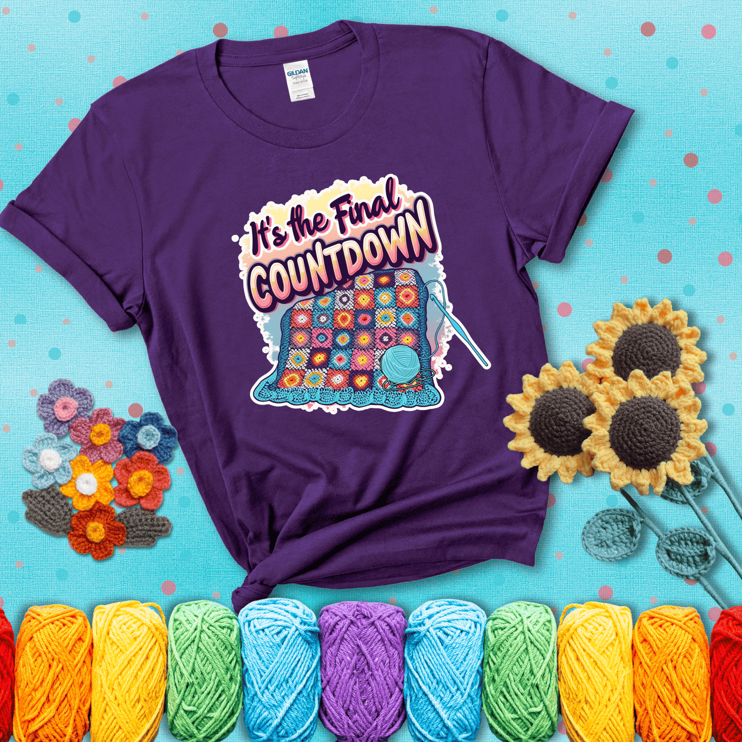 Colorful crochet blanket in progress with text "It’s the Final Countdown" above it in bold, multicolor lettering. A blue ball of yarn and hook are placed at the bottom, showing a purple tee with a teal sunflower background
