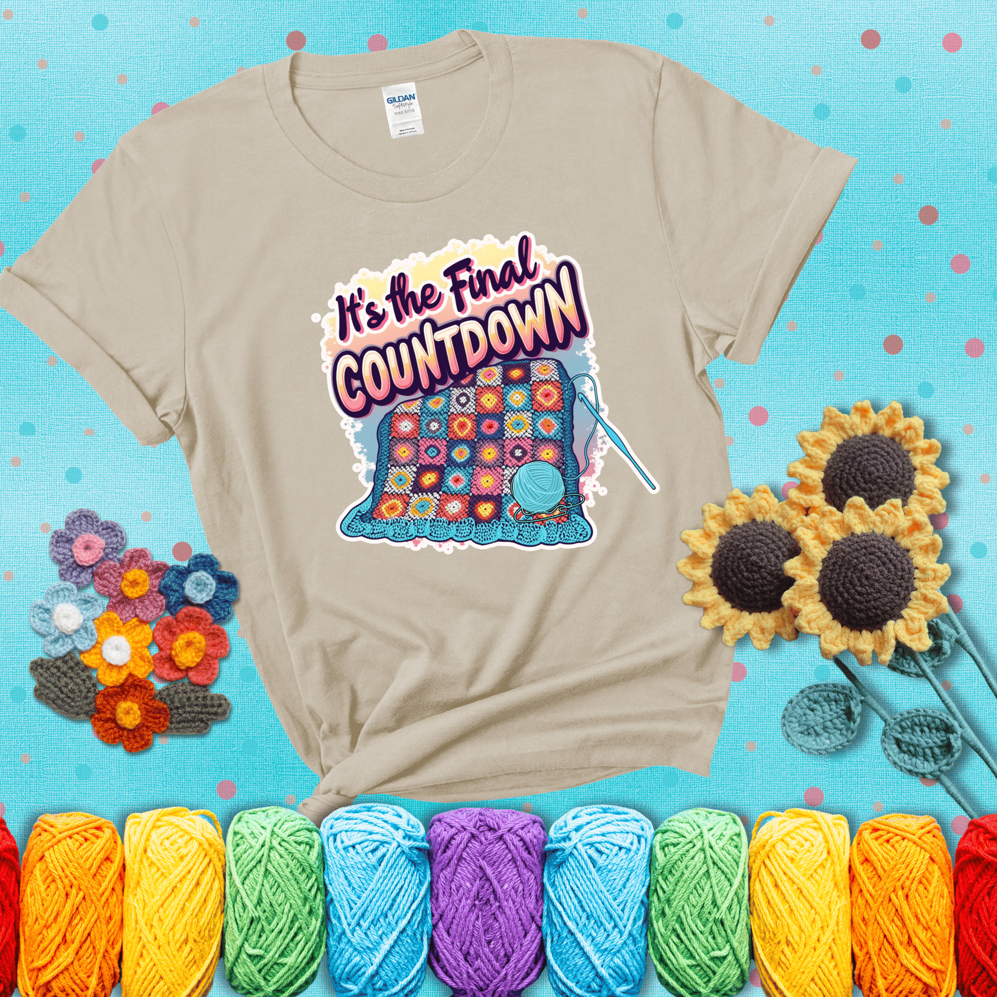 Colorful crochet blanket in progress with text "It’s the Final Countdown" above it in bold, multicolor lettering. A blue ball of yarn and hook are placed at the bottom, showing a beige tee with a teal sunflower background