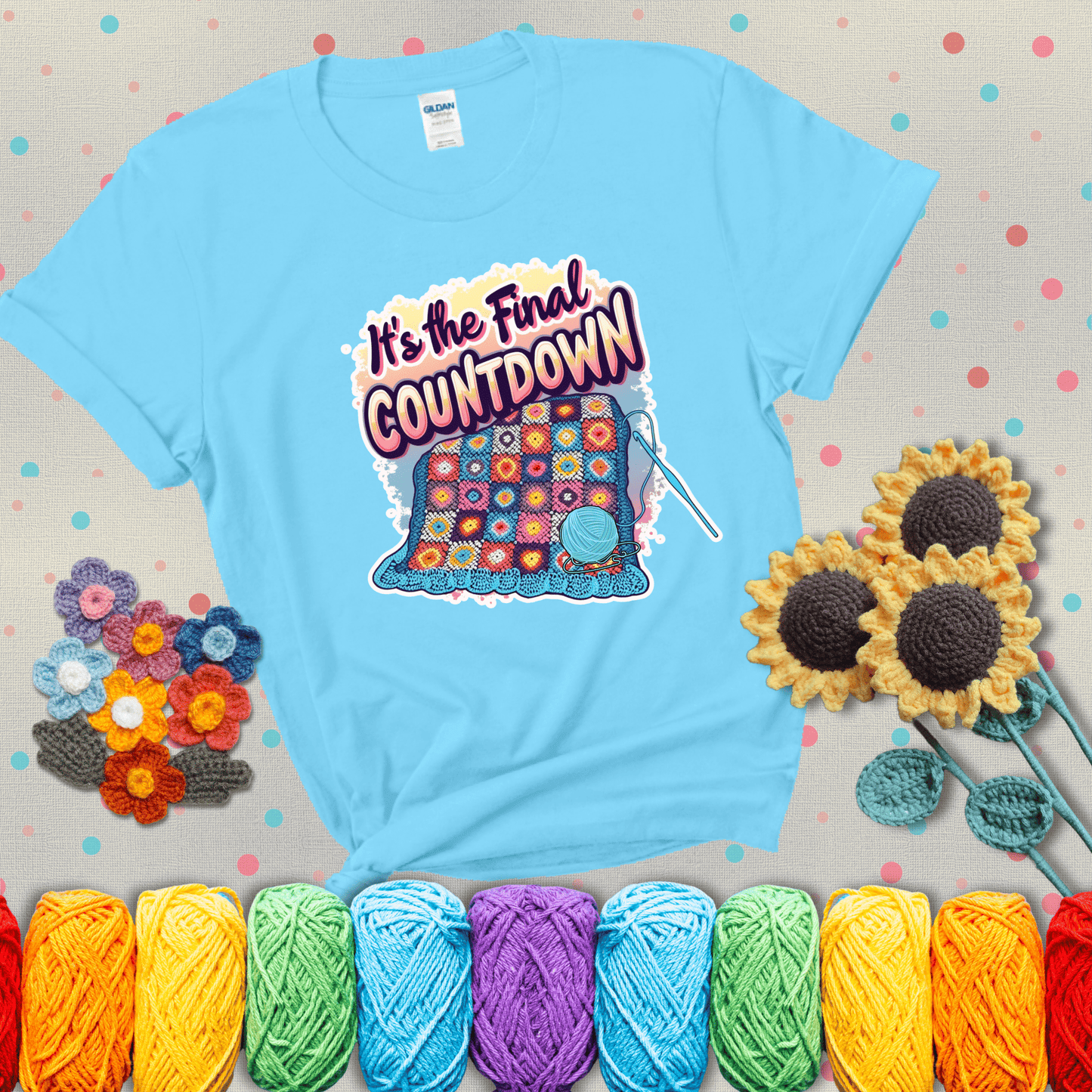 Colorful crochet blanket in progress with text "It’s the Final Countdown" above it in bold, multicolor lettering. A blue ball of yarn and hook are placed at the bottom, showing a light blue tee with grey sunflower background