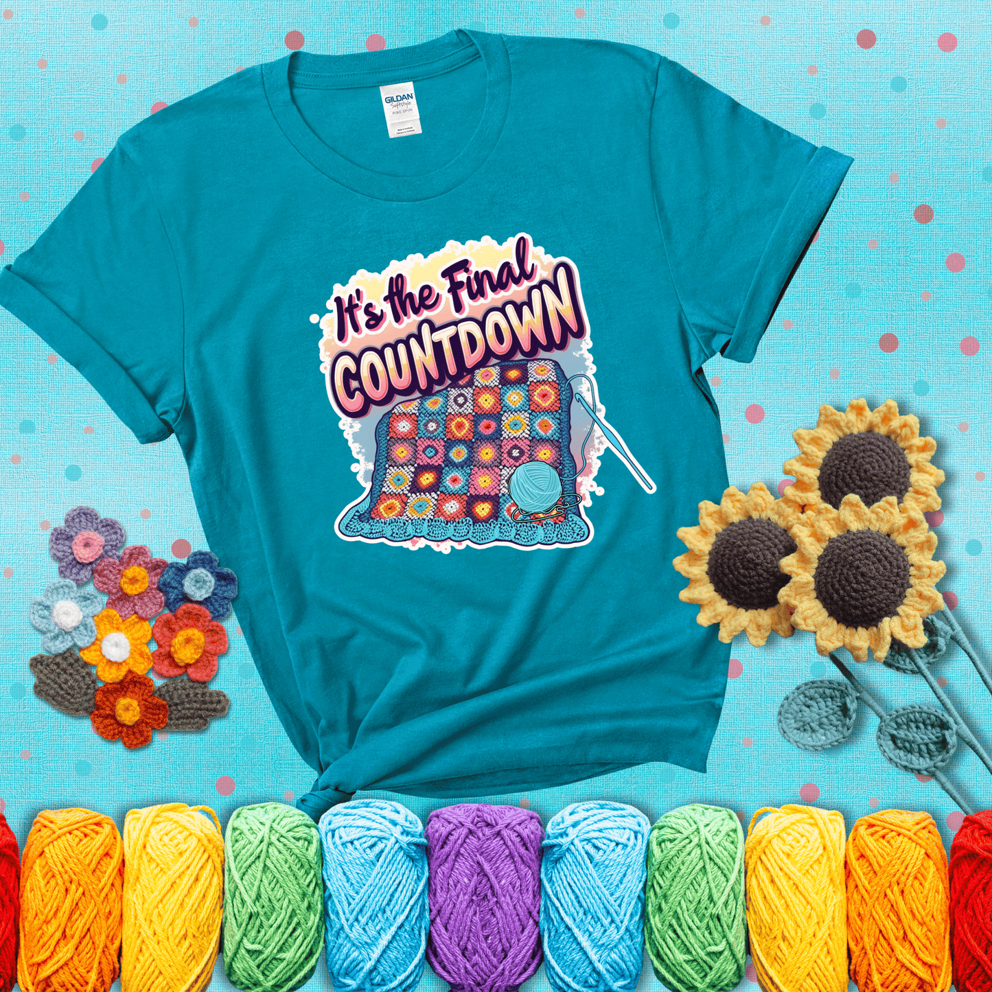 Colorful crochet blanket in progress with text "It’s the Final Countdown" above it in bold, multicolor lettering. A blue ball of yarn and hook are placed at the bottom, showing a blue-green tee with a teal sunflower background