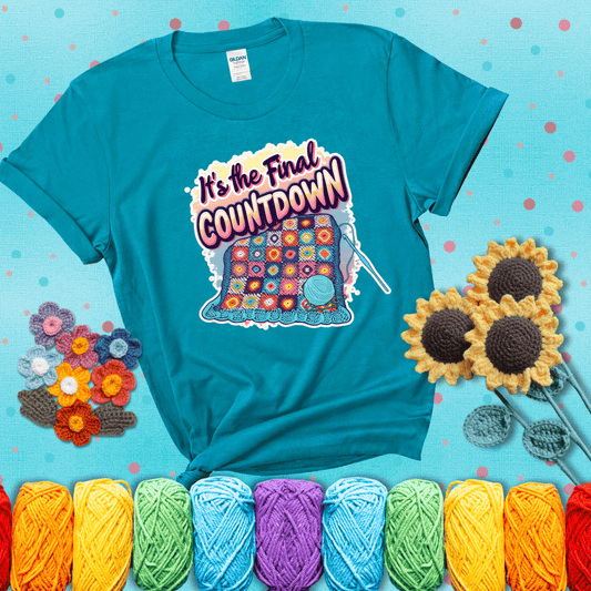 Colorful crochet blanket in progress with text "It’s the Final Countdown" above it in bold, multicolor lettering. A blue ball of yarn and hook are placed at the bottom, showing a blue-green tee with a teal sunflower background