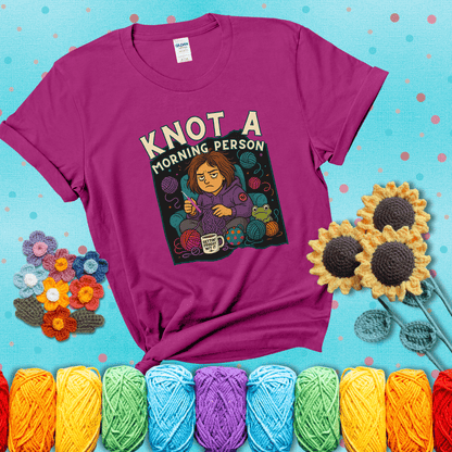 Sleepy crocheter in a purple hoodie sits in a chair with tangled yarn, coffee mug, and a frog sidekick, surrounded by yarn balls showing a magenta tee with a teal sunflower background