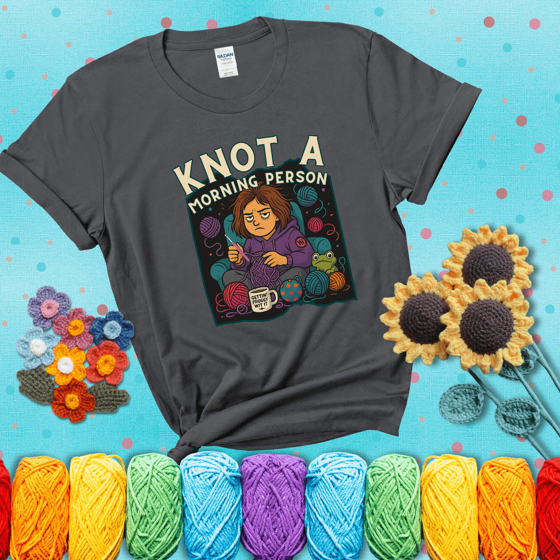 Sleepy crocheter in a purple hoodie sits in a chair with tangled yarn, coffee mug, and a frog sidekick, surrounded by yarn balls showing a dark grey tee with a teal sunflower background