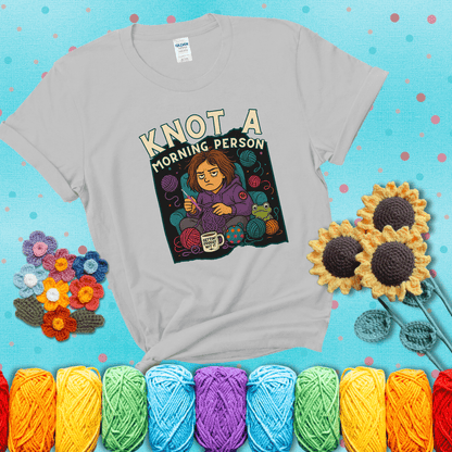 Sleepy crocheter in a purple hoodie sits in a chair with tangled yarn, coffee mug, and a frog sidekick, surrounded by yarn balls showing a light grey tee with a teal sunflower background