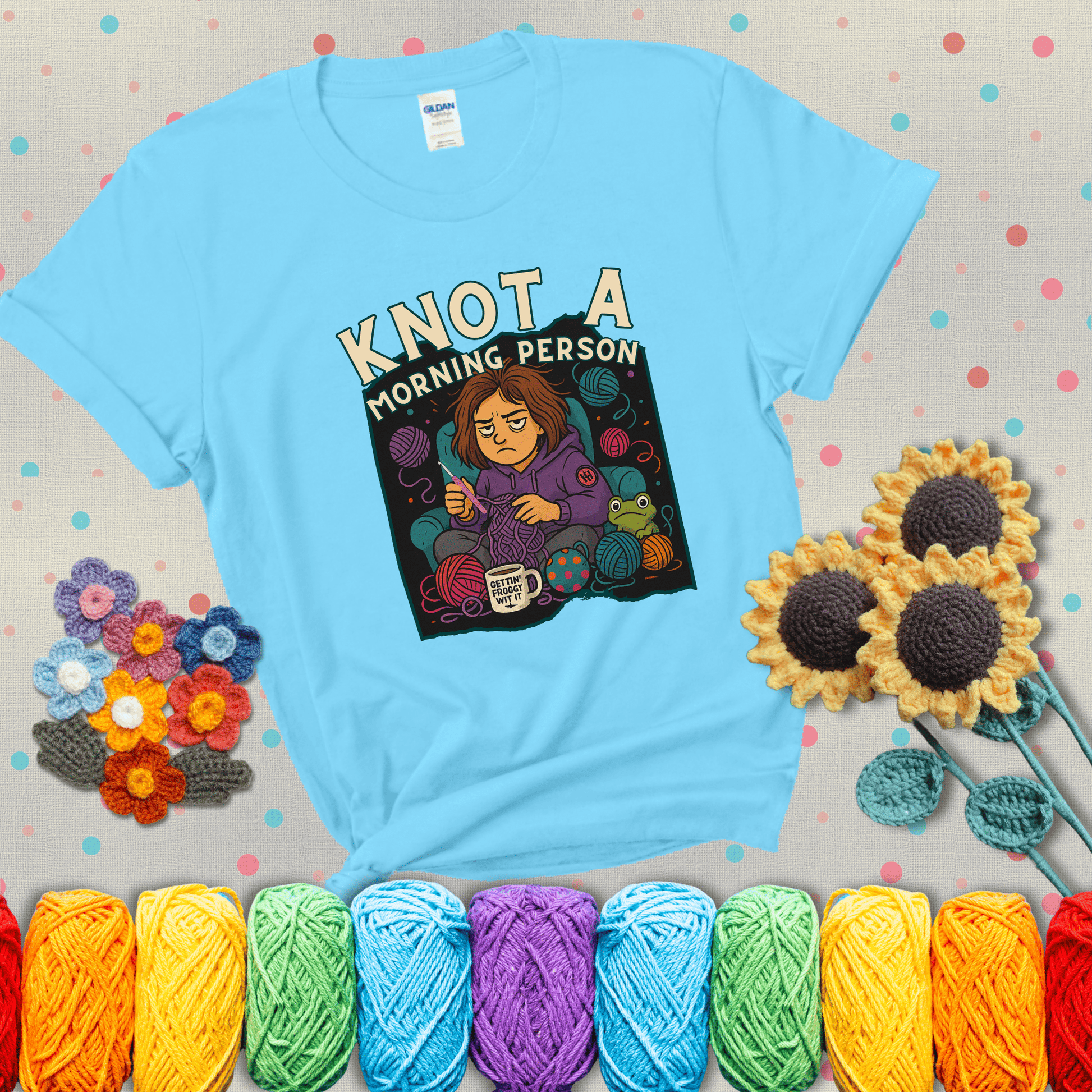 Sleepy crocheter in a purple hoodie sits in a chair with tangled yarn, coffee mug, and a frog sidekick, surrounded by yarn balls showing a light blue tee with grey sunflower background