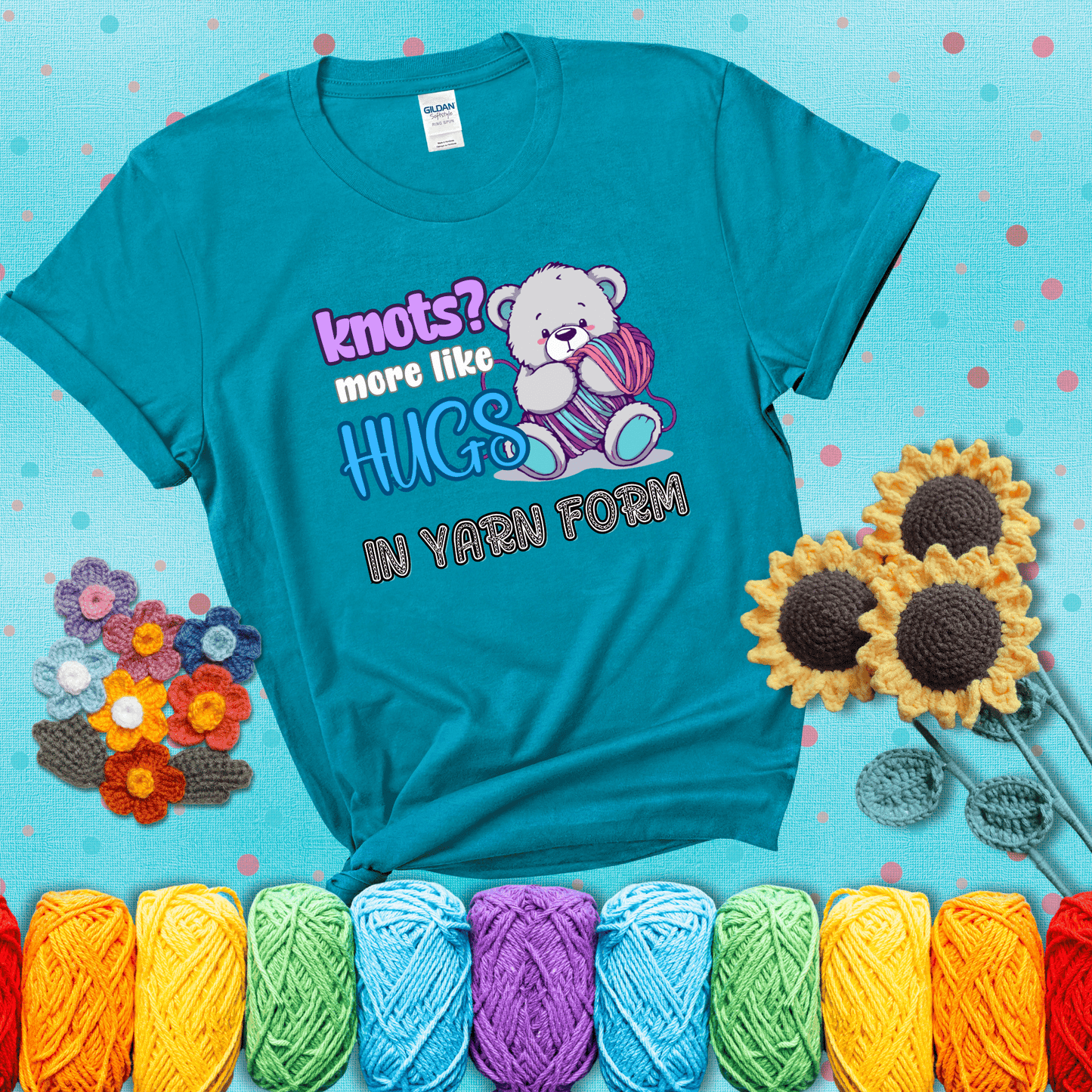 Cute teddy bear holding colorful yarn ball with hearts. Text reads: “Knots? More Like Hugs in Yarn Form.” Perfect for cozy gift-givers and yarn lovers, showing a blue-green tee with a teal sunflower background